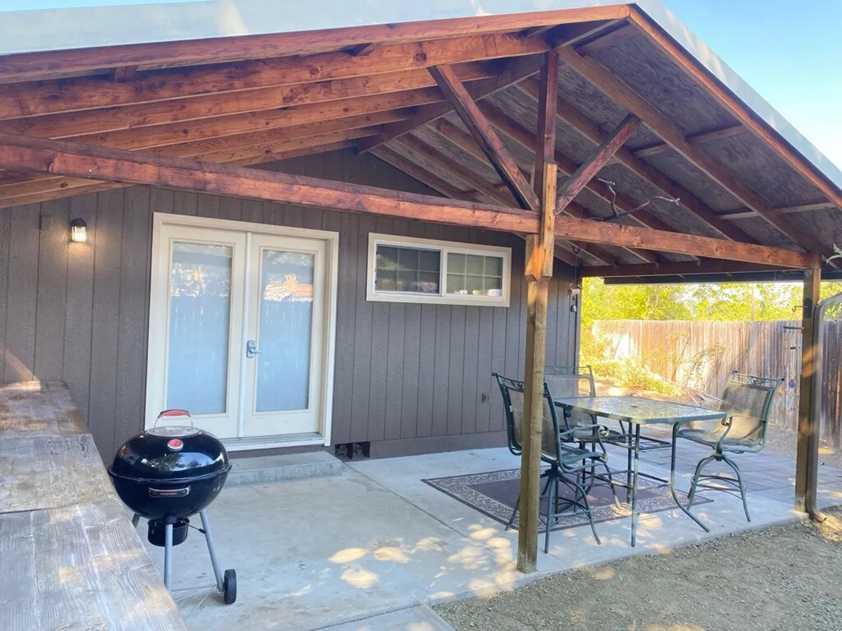 This image showcases a wooden patio structure that provides shade for an outdoor seating area, paired with a grill. Ample space is evident for groups to gather, relax, or enjoy meals. The setup is designed for barbecuing and evening entertainment, with comfortable seating and a welcoming, open layout that encourages social interaction.