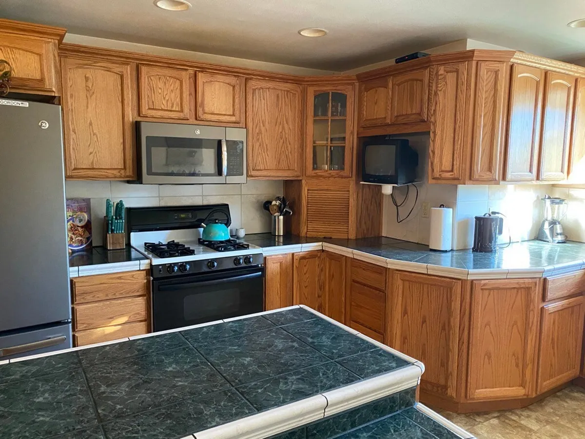 This image features a spacious kitchen with wooden cabinets, black appliances, and a large kitchen island. The teal kettle adds a pop of color, and the setup evokes a sense of comfort and homeliness. The space is clearly designed for cooking, gathering, and shared experiences.