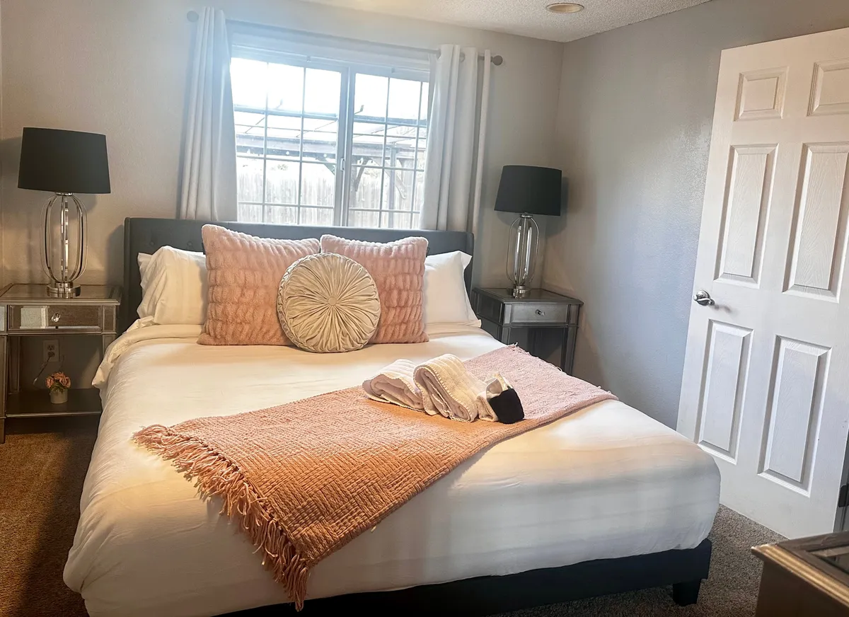 A cozy bedroom featuring a neatly made bed with decorative pillows, nightstands topped with lamps on either side, and a window that allows natural light to fill the space.