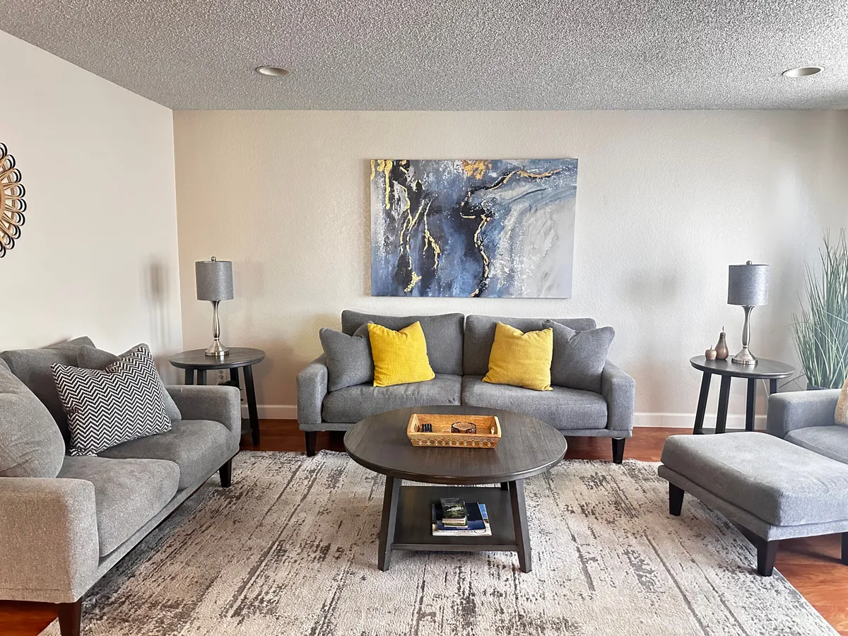 The image showcases a cozy, light-filled living room featuring plush sofas with cheerful yellow cushions, a stylish coffee table at the center, and an eye-catching abstract painting on the wall. The space is designed for relaxation and social gatherings, inviting guests to unwind, play games, or enjoy coffee mornings together.
