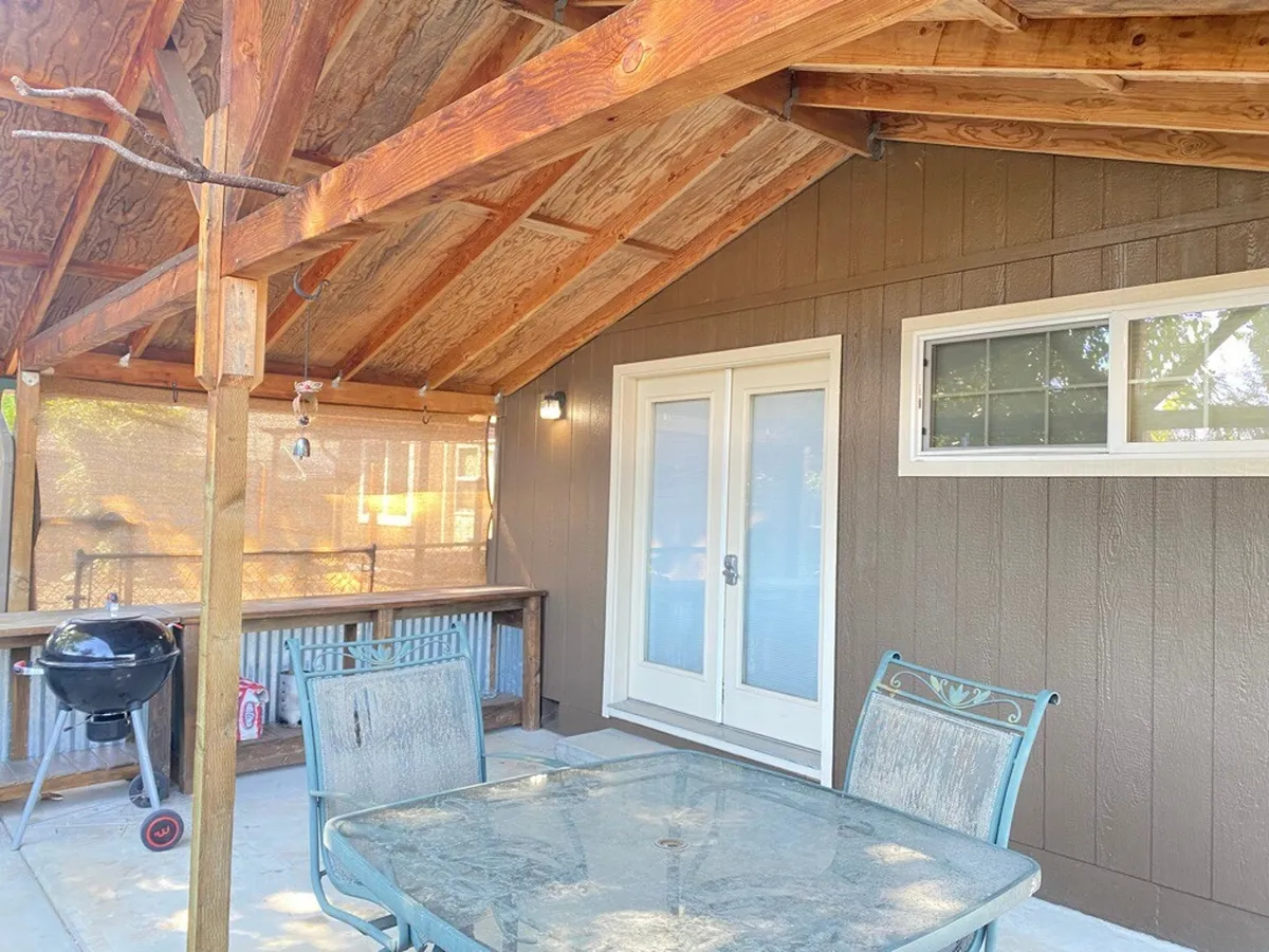 This image showcases a cozy outdoor dining area under wooden beams, with a dining table set for gatherings, sliding glass doors connecting to the home's interior, and a barbecue grill ready for family cookouts.