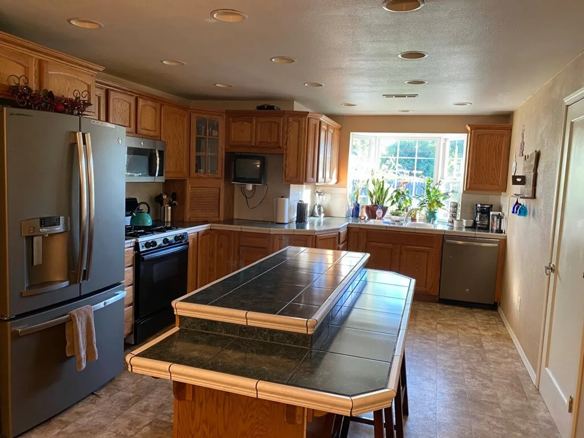 A warm and welcoming kitchen scene with a prominent island, window with plants, refrigerator, and stove. The open layout invites group cooking, conversation, and shared mealtimes.