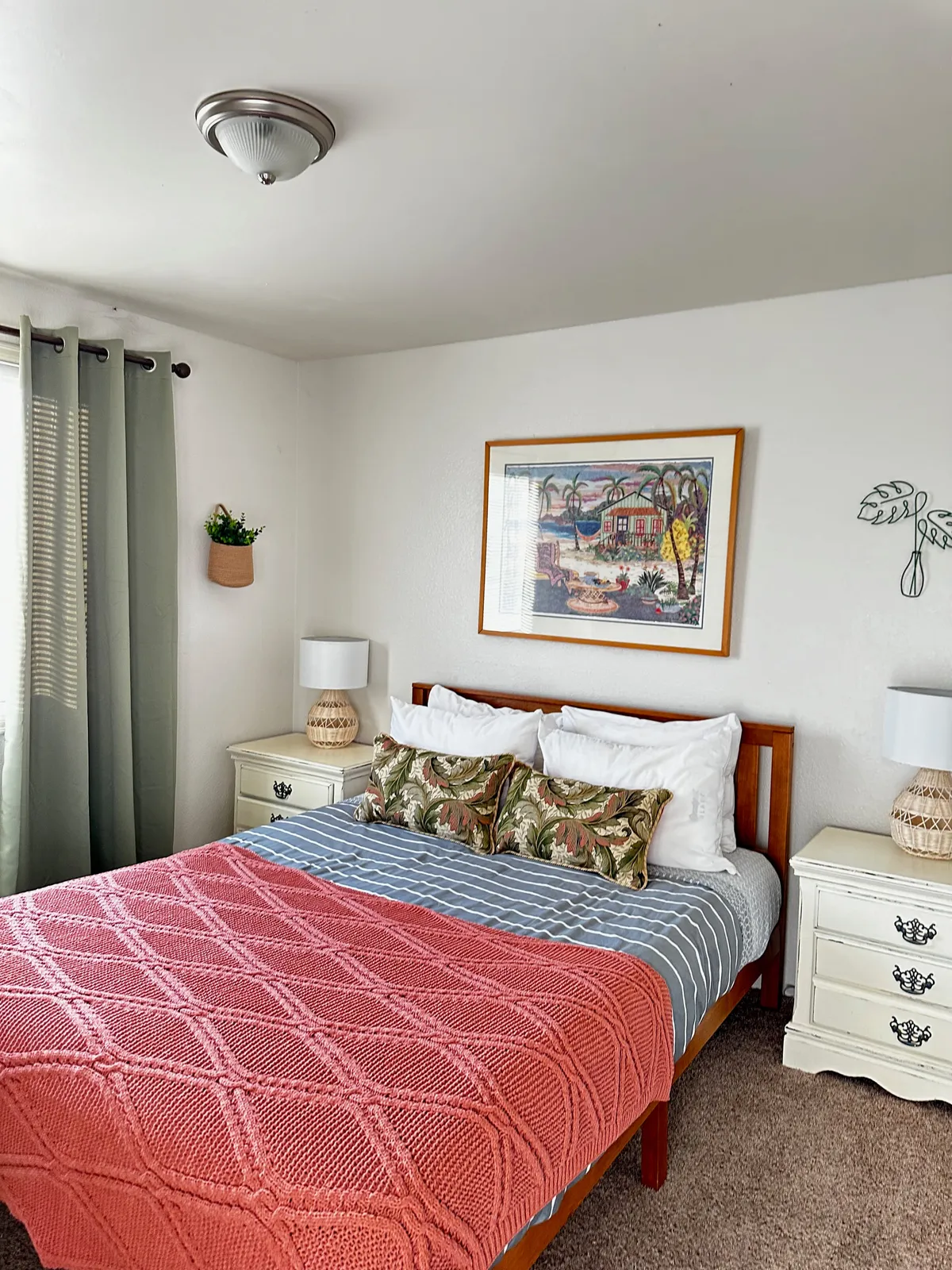 A tranquil bedroom scene with a bed draped in a pink knitted blanket, framed artwork above the headboard, matching nightstands with lamps, and soft curtains framing the window.