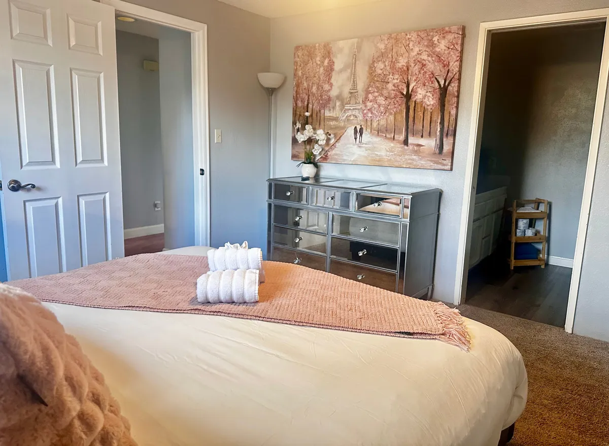 This image showcases a plush bed draped with blankets and towels, a decorative painting of a Parisian street above the headboard, and a mirrored dresser reflecting soft light. The room feels serene and inviting, ideal for unwinding after a busy day.